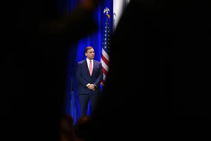 A politician stands on a stage, with his hands folded in front of him. The view of the of the stage is silhouetted by the shoulders of onlookers in the crowd.